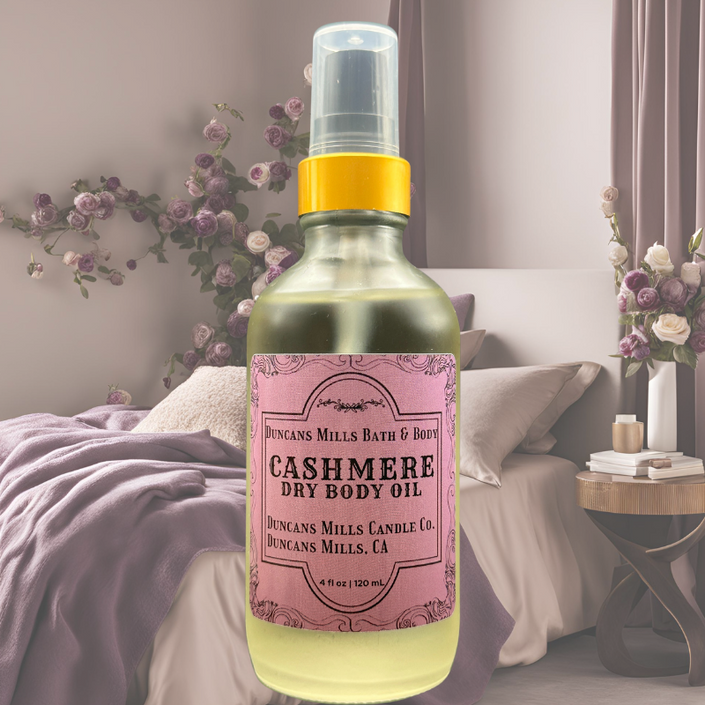Cashmere Body Oil is displayed with a bedroom background of flowers and cozy plum blankets. 