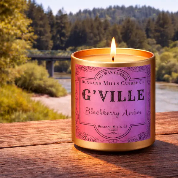 Candle with 'G'Ville' label on a wooden surface near the Russian River and redwood trees.