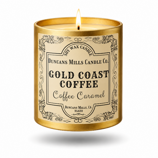 A lit scented candle in a cylindrical gold container with a cream label reading 'Duncans Mills Candle Co. Gold Coast Coffee Coffee Caramel' on a white background.
