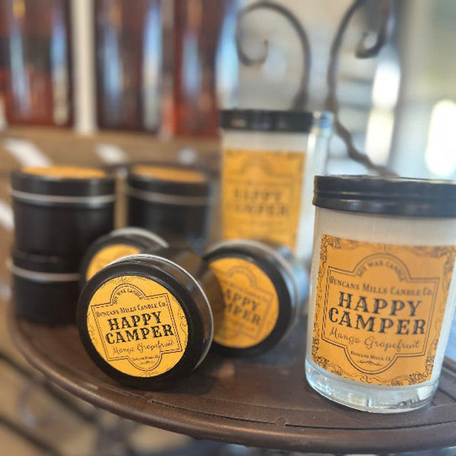 Display of several lidded candles of two styles with a yellow label: Happy Camper, Mango Grapefruit