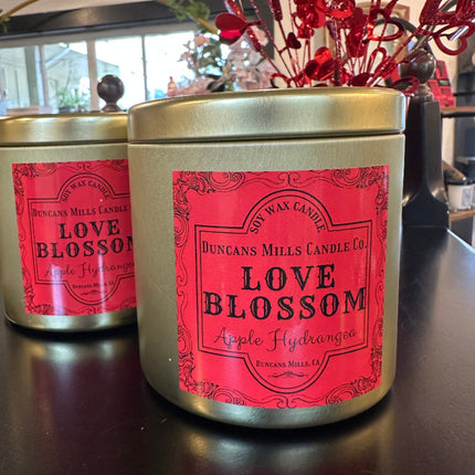 Large gold-colored candle tins with lids and bright reddish-pink label: Love Blossom, Apple Hydrangea