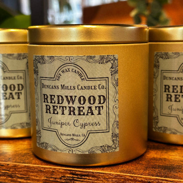 Large gold-colored candle tins with lids and pale-green label: Redwood Retreat, Juniper Cypress