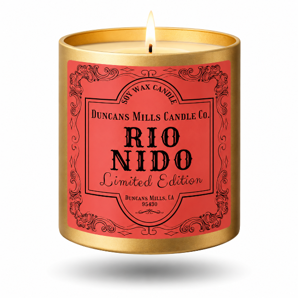 A lit scented soy wax candle in a cylindrical gold metal container with a coral-red label. The label features black decorative borders and text reading 'Duncans Mills Candle Co.