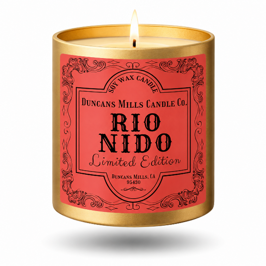 A lit scented soy wax candle in a cylindrical gold metal container with a coral-red label. The label features black decorative borders and text reading 'Duncans Mills Candle Co.
