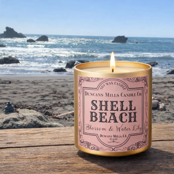 Candle labeled 'Shell Beach' by Duncan's Mills Candle Co. on a beach with ocean view.