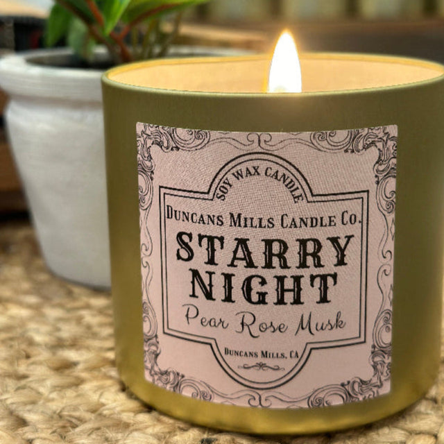 Large gold-colored candle tin with candle lit, lid not shown, and pale-pink label: Starry Night, Pear Rose Musk