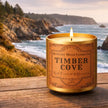 Candle labeled 'Timber Cove' by Duncan's Mills Candle Co. on a wooden surface with a scenic ocean background.