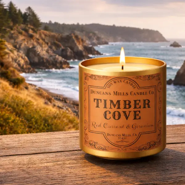 Candle labeled 'Timber Cove' by Duncan's Mills Candle Co. on a wooden surface with a scenic ocean background.